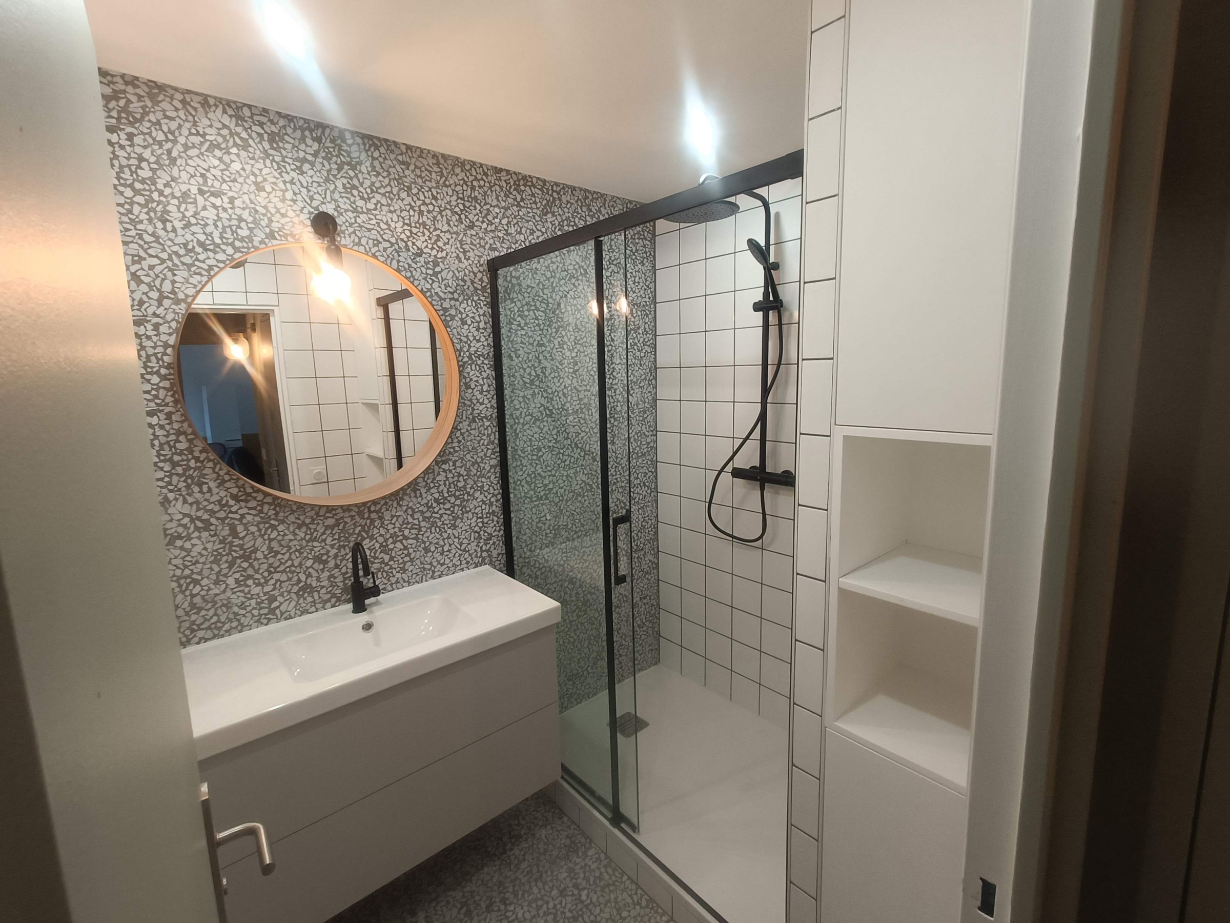 Salle de bains avec miroir rond, meuble vasque gris et douche vitrée à montants noirs